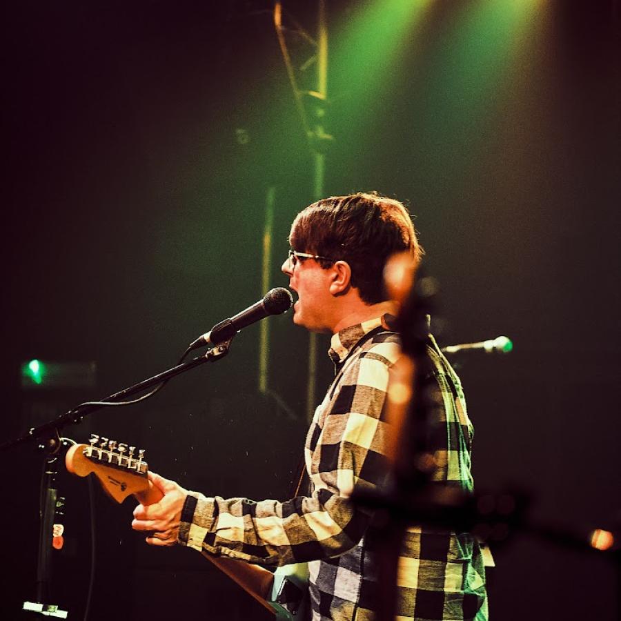 A man wearing a checked shirt is playing a Fender Jazzmaster guitar and singing into a microphone. The background is blurred with two spotlights behind him. 